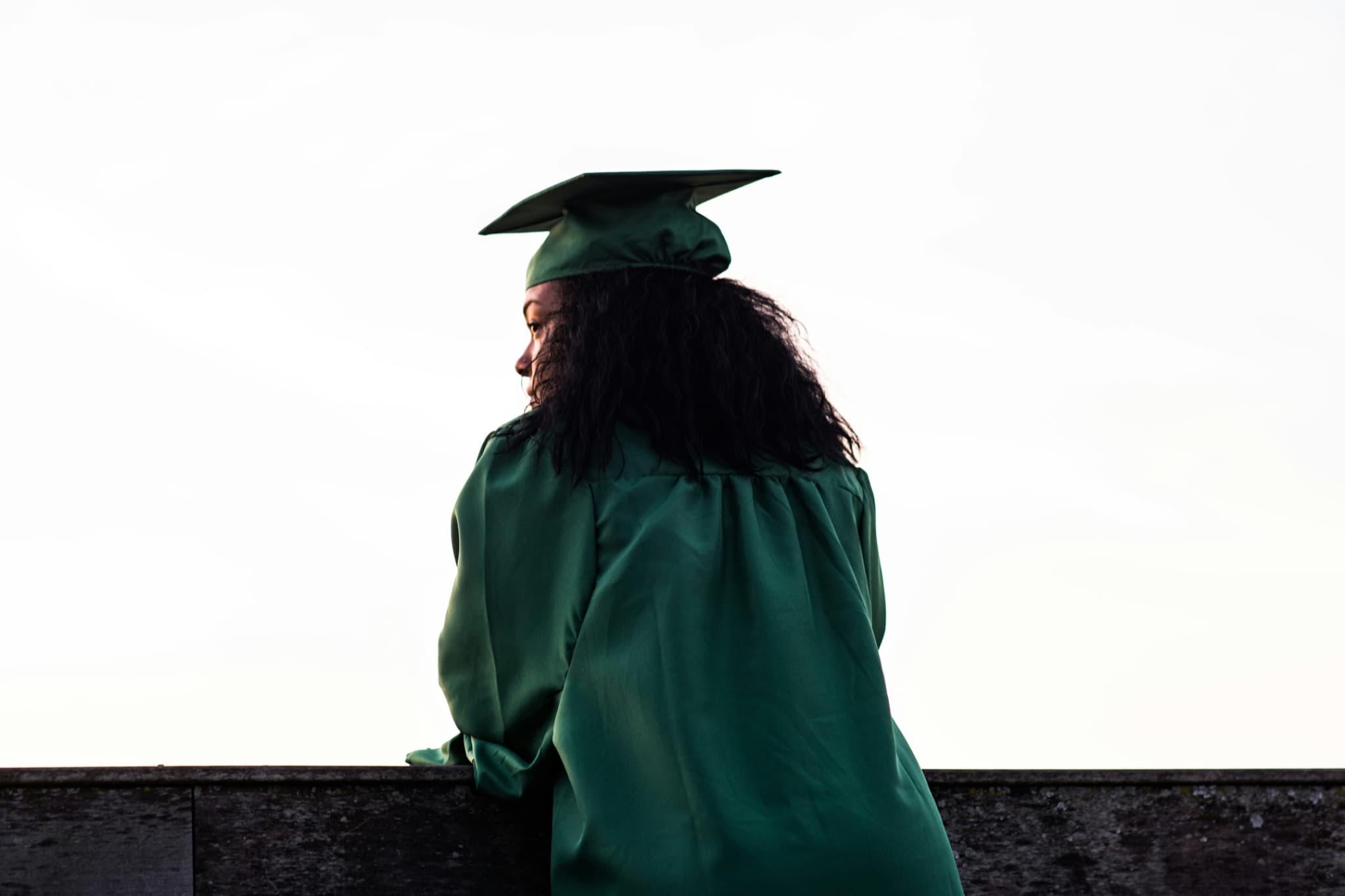 A graduate in cap and gown, seen from behind at a parapet, head turned to one side against a bright sky