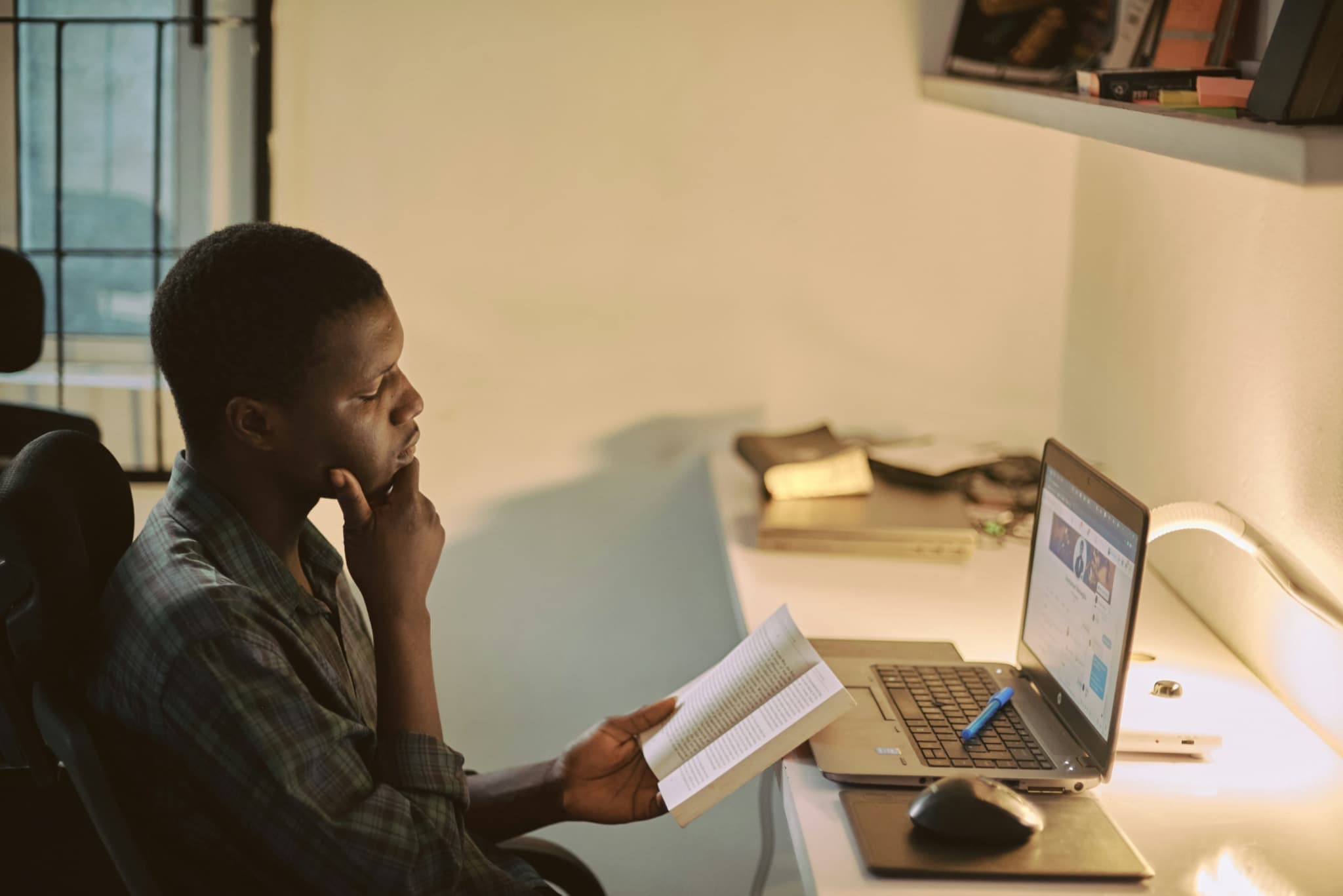 A student at a desk in the evening, holding an open book and looking at a laptop screen