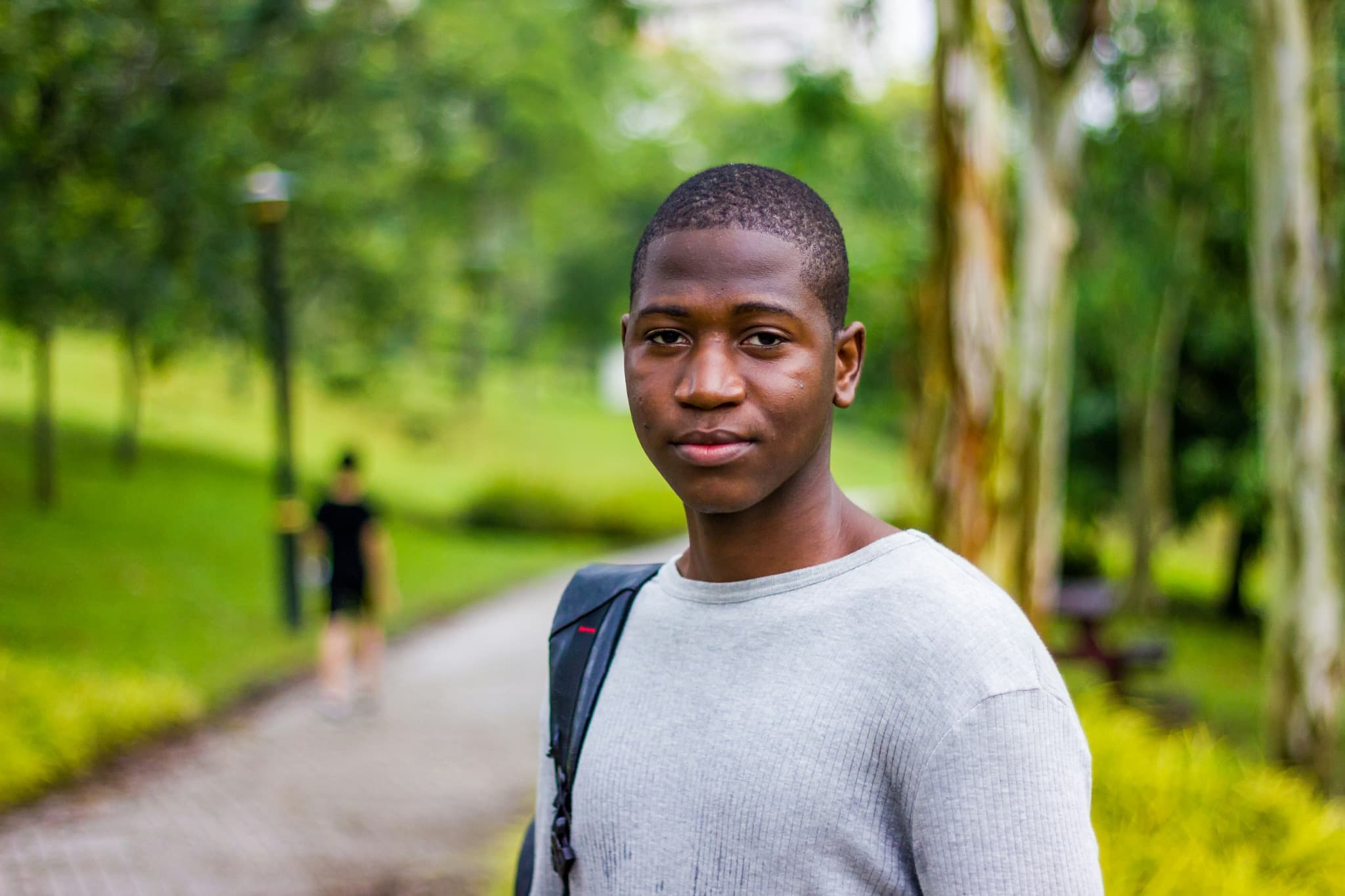 A student with a backpack standing on a tree-lined campus path, looking towards the camera